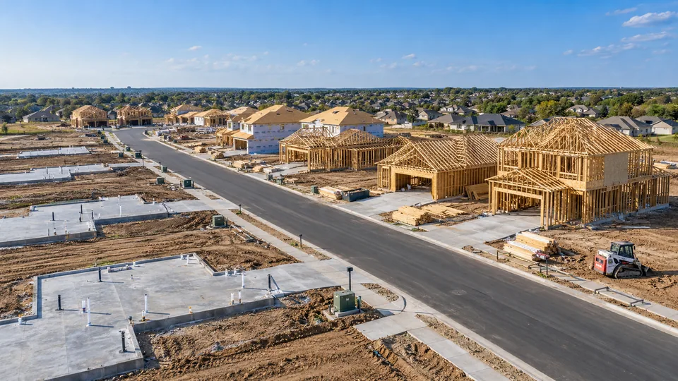 Aerial view of a suburban tract under construction, with finished slabs and plumbing stub-ups on one side of a new street, framed homes on the other and a compact track loader beside stacked lumber